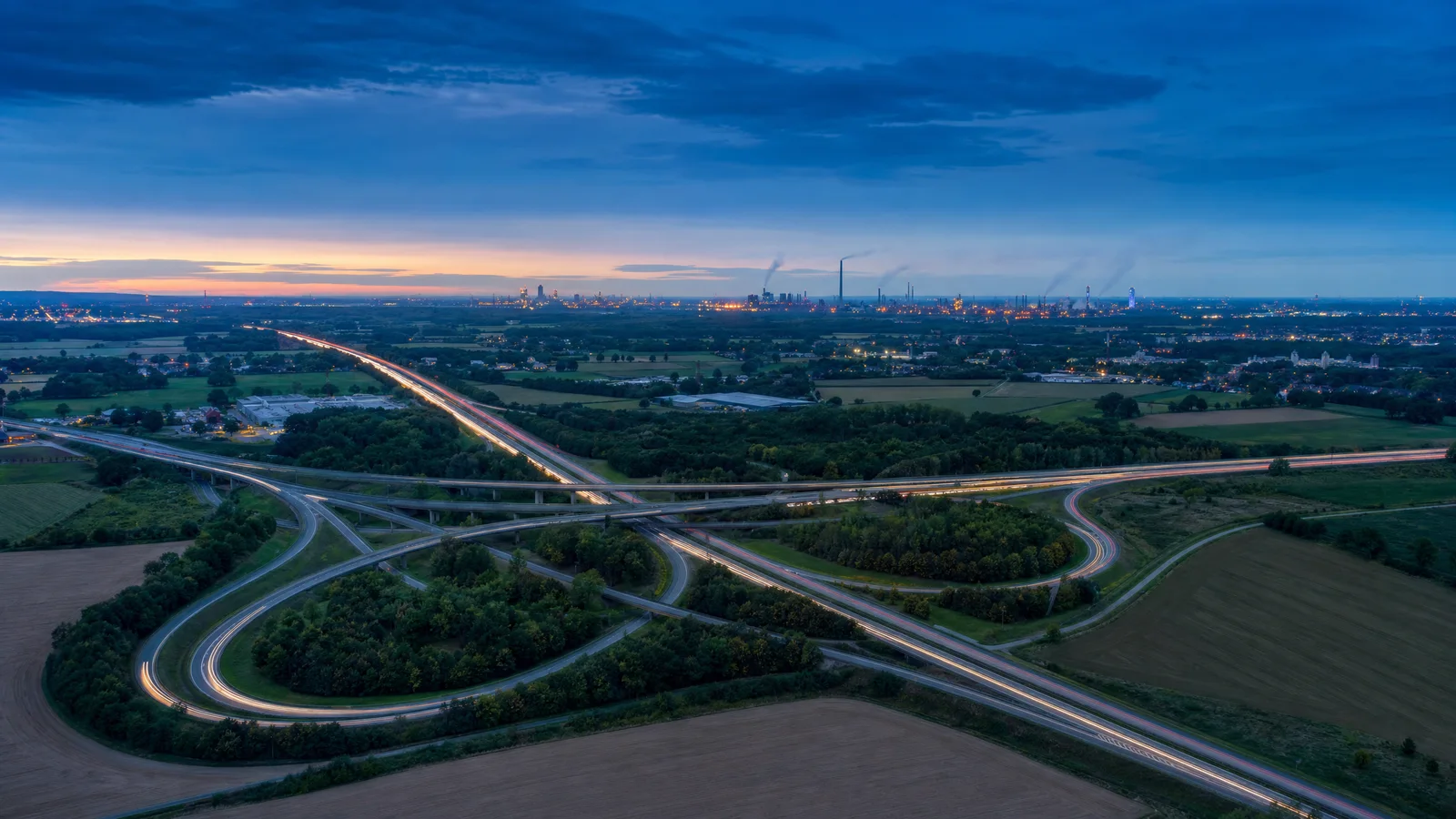 Luftaufnahme eines Autobahnkreuzes im Münsterland zur blauen Stunde, im Hintergrund die Industriesilhouette des Ruhrgebiets